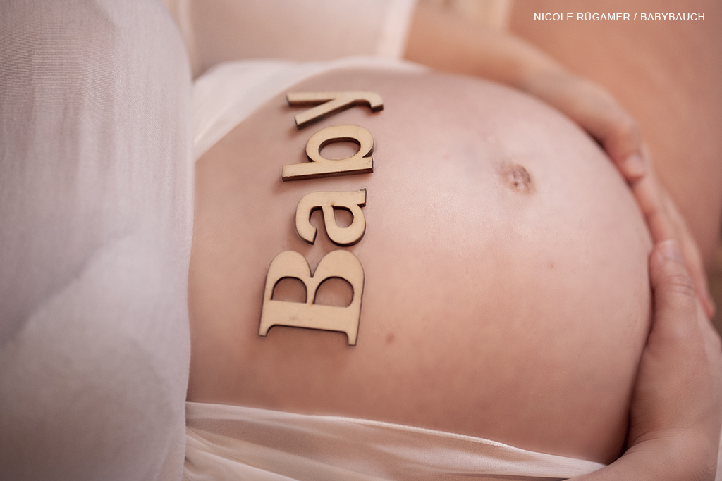auf dem Babybauch liegt mit Holzbuchstaben das Wort Baby